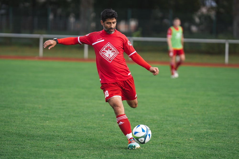 homme en maillot de football rouge donnant un coup de pied dans un ballon de football sur un terrain d’herbe verte pendant la journée