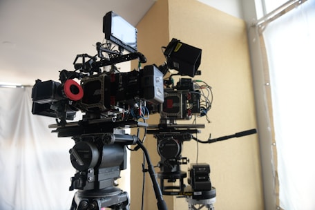 Two professional film cameras are mounted on tripods in a well-lit room. The cameras have various attachments, including monitors, cables, and other accessories. The background features a large window with white curtains and a beige-colored wall.