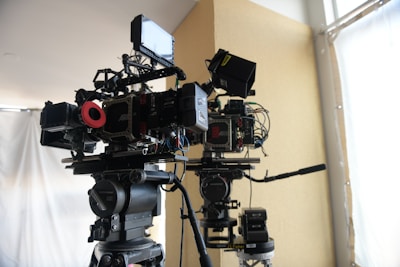 Two professional film cameras are mounted on tripods in a well-lit room. The cameras have various attachments, including monitors, cables, and other accessories. The background features a large window with white curtains and a beige-colored wall.