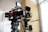 Two professional film cameras are mounted on tripods in a well-lit room. The cameras have various attachments, including monitors, cables, and other accessories. The background features a large window with white curtains and a beige-colored wall.