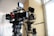 Two professional film cameras are mounted on tripods in a well-lit room. The cameras have various attachments, including monitors, cables, and other accessories. The background features a large window with white curtains and a beige-colored wall.