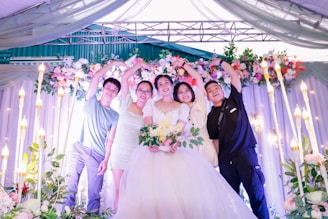 A group of five people stand under an arch decorated with white and pink flowers. They are smiling and have their arms raised in celebration. The central figure is wearing a white wedding dress and holding a bouquet, suggesting a wedding ceremony.