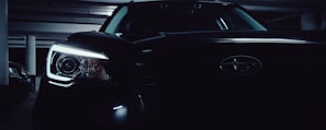 A close-up view of the front of a black car in a dimly lit parking garage, focusing on the headlight and the logo on the grille.