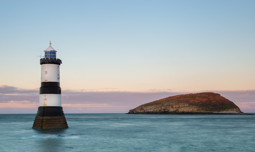 Phare blanc et noir sur une formation rocheuse brune près d’un plan d’eau pendant la journée