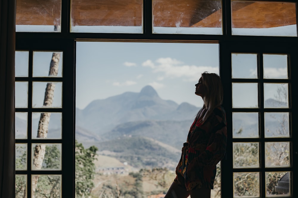 赤と黒の格子縞のドレスシャツを着た女性が窓辺に立って昼間、山を眺めている