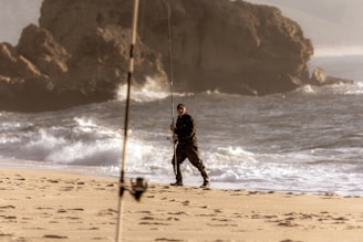 A person holding a fishing rod stands on a sandy beach near the shoreline, with waves crashing in the background and large rocky formations in the distance. Another fishing rod is visible in the foreground, slightly out of focus.