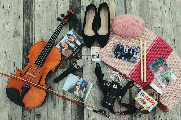 A violin with its bow lies on a wooden surface surrounded by various items, including a pair of black shoes, pink yarn, and knitting needles placed on a knitted scarf. Several photographs are scattered around, along with a microphone, camera, and a jar of Nutella.