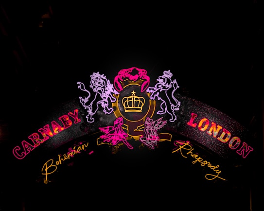 Neon signs featuring the words 'Carnaby' and 'London' in bold red, with additional handwritten-style text of 'Bohemian' and 'Rhapsody' in yellow. Outlined figures of lions and a crown are illuminated in bright colors against a dark background.