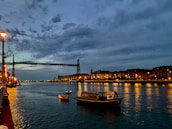 A calm river with boats passing under a city bridge at sunset.