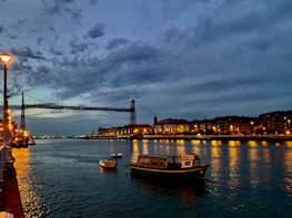 A calm river with boats passing under a city bridge at sunset.