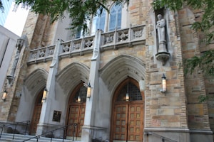 A grand entrance of a stone building featuring three large arched wooden doors, flanked by intricate columns and lanterns. Above the doors are detailed stone carvings, including statues and ornamental designs. The facade showcases a blend of buff and tan stone, contributing to the historic and architectural elegance.