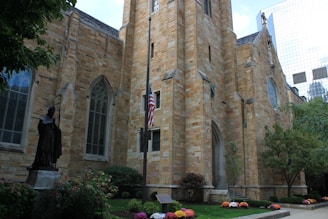 A large, historic stone church with gothic architecture featuring tall arched windows, surrounded by neatly maintained bushes and flowers. An American flag is hoisted on a flagpole in front of the church alongside a statue of a robed figure. The scene is set against a backdrop of trees and a modern glass building on the right.