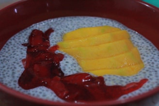 A bowl containing chia pudding is adorned with vibrant yellow mango slices and a drizzle of dark red berry sauce. The black chia seeds create a speckled texture against the light pudding base, providing a contrast to the smoothness of the mango slices and the glossy surface of the berry sauce.