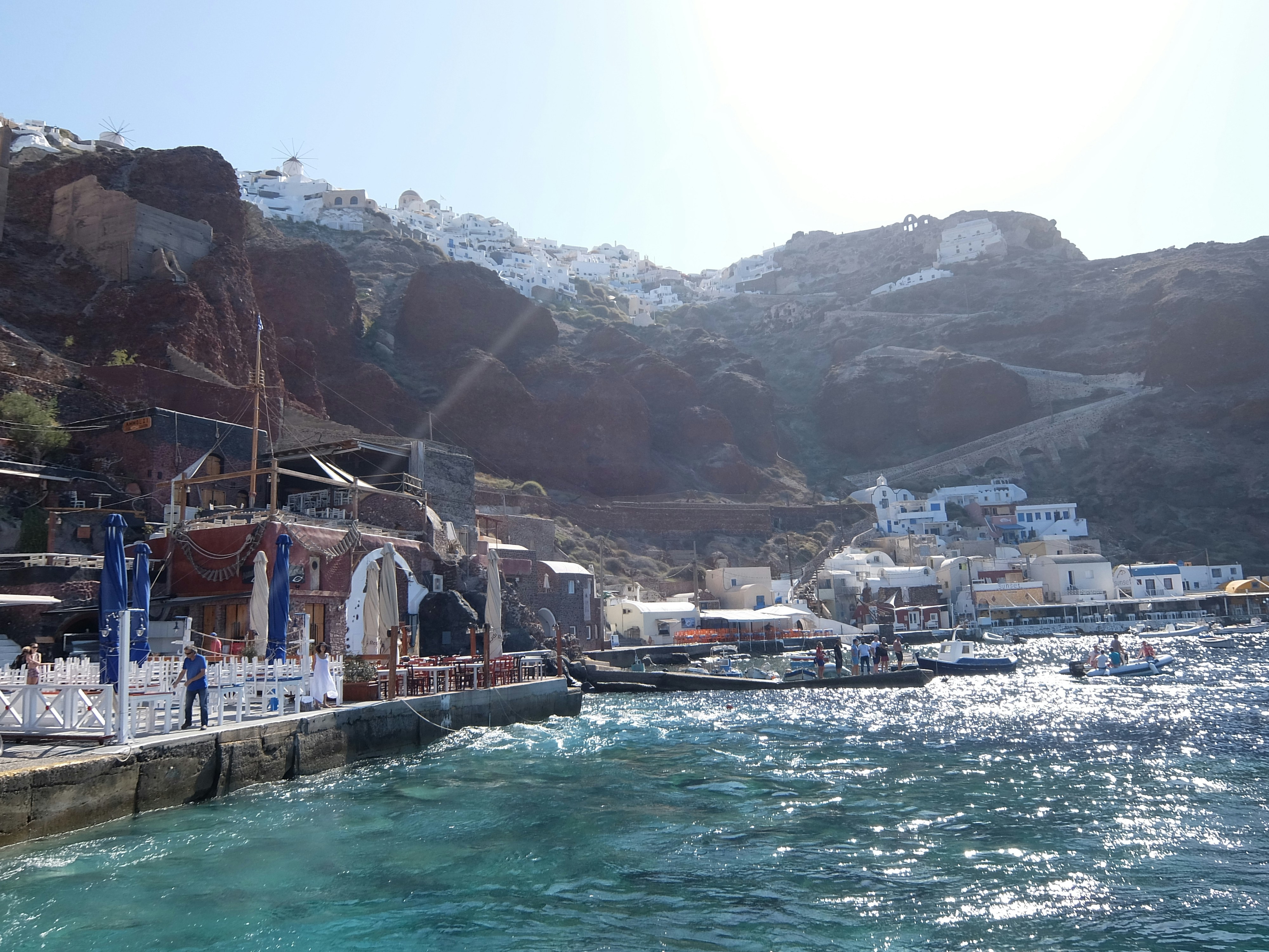 White-washed Santorini buildings cascading down volcanic cliffs, deep blue Aegean sea, bright midday Mediterranean light