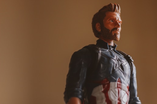 A toy figure of a superhero dressed in a blue, red, and white costume with a star emblem on the chest is posed against a plain beige background. The figure has a prominent beard and a serious expression.