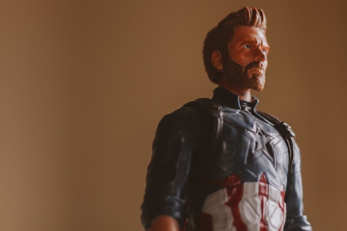 A toy figure of a superhero dressed in a blue, red, and white costume with a star emblem on the chest is posed against a plain beige background. The figure has a prominent beard and a serious expression.