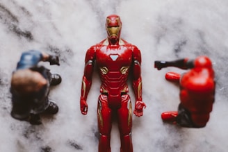 Three action figures are placed on a marble-like surface. The central figure is in a red and gold armored suit, laying flat on its back. Two other figures with darker outfits are positioned on either side, partially out of focus.