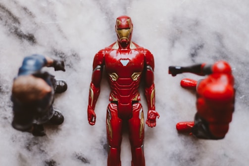 Three action figures are placed on a marble-like surface. The central figure is in a red and gold armored suit, laying flat on its back. Two other figures with darker outfits are positioned on either side, partially out of focus.