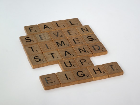 Wooden Scrabble tiles arranged on a white background, displaying words such as 'FALL', 'SEVEN', 'TIMES', 'STAND', 'UP', and 'EIGHT'. The tiles are placed in a grid-like formation.