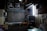 A dimly lit, cluttered room with industrial and storage elements. There are two air conditioning units mounted on the upper wall, a rolled-up shutter door in the center, and various tables and shelves scattered around. Exposed wiring and a bright fluorescent light tube are visible, while some parts of the room appear dusty and unused.