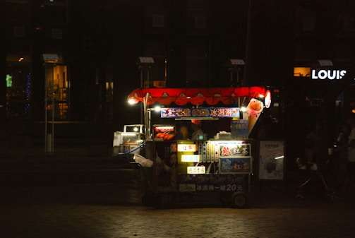 A brightly lit street food cart is situated in a dimly lit urban environment. The cart has a red canopy with various illuminated signs displaying menu items. The surrounding area is dark, highlighting the cart as the focal point.