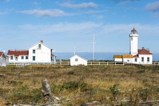 A coastal landscape featuring a lighthouse with a red roof, accompanied by several white buildings with matching roofs. The foreground consists of grassy fields with scattered driftwood, bordered by a simple white fence. The sky is partly cloudy, casting a serene atmosphere over the scene.
