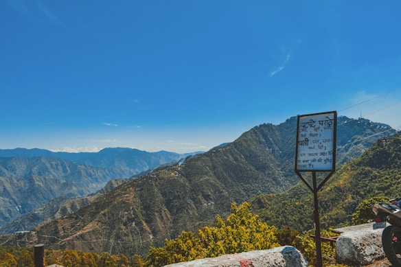 A scenic mountainous landscape with lush greenery under a clear blue sky. There is a roadside sign in the foreground with text and cable lines visible. A motorbike can be partially seen on the right.