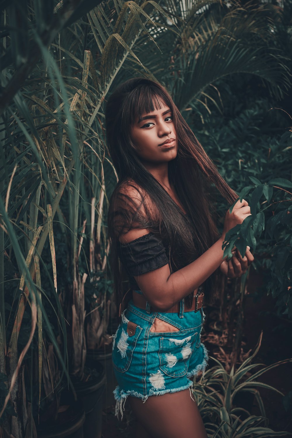woman in black crop top and blue denim shorts standing beside green plants