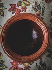A large, rustic clay pot is positioned centrally, exhibiting a deep brown color with a glossy finish. The pot sits on a floral-patterned surface with vibrant designs of leaves, pears, and small fruits, adding an autumnal feel to the composition.