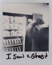 A blurred black and white photograph featuring a vending machine partially visible on the left and an indistinct figure on the right, creating a mysterious and ghostly effect. The text 'I Saw a Ghost' is handwritten below the image.