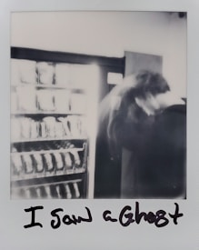 A blurred black and white photograph featuring a vending machine partially visible on the left and an indistinct figure on the right, creating a mysterious and ghostly effect. The text 'I Saw a Ghost' is handwritten below the image.