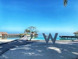 A scenic view of a resort with overwater bungalows stretching over a calm turquoise sea. A large decorative letter 'W' stands on a wooden walkway in the foreground. There is a small tree and clear blue sky, with palm leaves slightly visible in the corner.