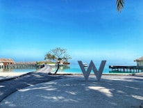 A scenic view of a resort with overwater bungalows stretching over a calm turquoise sea. A large decorative letter 'W' stands on a wooden walkway in the foreground. There is a small tree and clear blue sky, with palm leaves slightly visible in the corner.