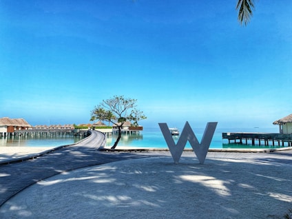 A scenic view of a resort with overwater bungalows stretching over a calm turquoise sea. A large decorative letter 'W' stands on a wooden walkway in the foreground. There is a small tree and clear blue sky, with palm leaves slightly visible in the corner.