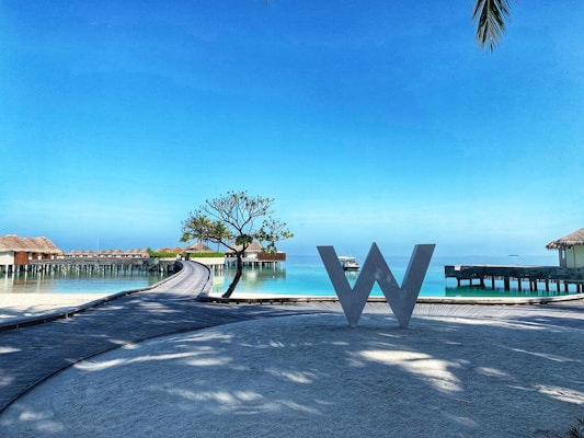 A scenic view of a resort with overwater bungalows stretching over a calm turquoise sea. A large decorative letter 'W' stands on a wooden walkway in the foreground. There is a small tree and clear blue sky, with palm leaves slightly visible in the corner.