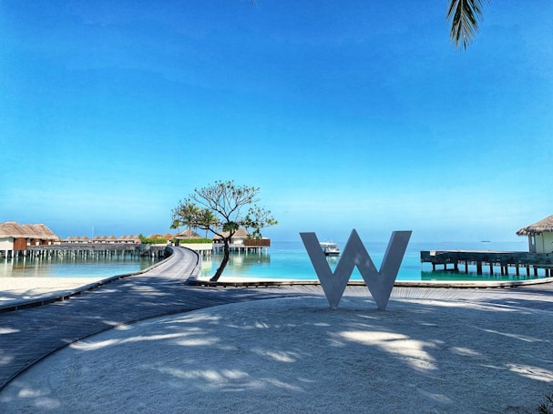 A scenic view of a resort with overwater bungalows stretching over a calm turquoise sea. A large decorative letter 'W' stands on a wooden walkway in the foreground. There is a small tree and clear blue sky, with palm leaves slightly visible in the corner.