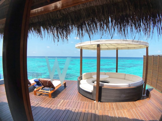 A luxurious outdoor seating area overlooks a vibrant turquoise ocean, featuring a round sofa under a thatched roof and a couple of sun loungers on a wooden deck. An idyllic and serene seascape is visible in the background.