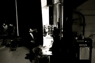A man in a suit stands backstage drinking from a bottle, with a dimly lit behind-the-scenes environment. In the background, an orchestra is visible on stage, illuminated by bright lights. The scene captures a contrast between the dark backstage area and the brightly lit stage.