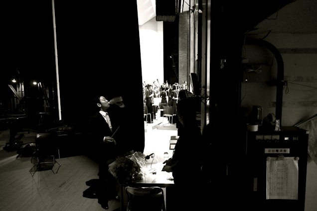 A man in a suit stands backstage drinking from a bottle, with a dimly lit behind-the-scenes environment. In the background, an orchestra is visible on stage, illuminated by bright lights. The scene captures a contrast between the dark backstage area and the brightly lit stage.