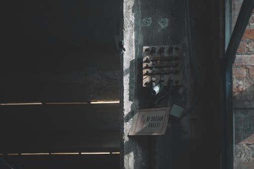 A dimly lit industrial space with a rough concrete wall, featuring an old electrical panel mounted on it. The panel has multiple wires and components, giving a sense of neglect or disuse. A warning sign with cautionary text is affixed below, with some cables dangling beside it. The surrounding area shows a mix of concrete and brick surfaces, with shadows emphasizing the aged and somewhat ominous atmosphere.