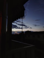 The view captures a tranquil evening scene from a balcony, silhouetted against a twilight sky. Dark clouds are scattered across the dimly lit sky, with a faint hint of the setting sun on the horizon. The balcony railing is visible in the foreground, and a building is obscured in the shadow.