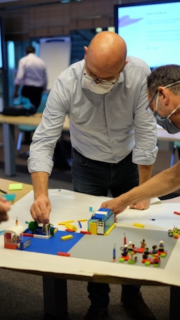 Two individuals are focused on constructing a model using colorful building blocks on a table. One person is bald and wearing glasses, both are dressed in business casual attire. The setting appears to be a workspace or meeting room with a screen and a flip chart in the background. Bright lighting highlights the scene, emphasizing a collaborative atmosphere.