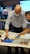 Two individuals are focused on constructing a model using colorful building blocks on a table. One person is bald and wearing glasses, both are dressed in business casual attire. The setting appears to be a workspace or meeting room with a screen and a flip chart in the background. Bright lighting highlights the scene, emphasizing a collaborative atmosphere.