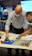 Two individuals are focused on constructing a model using colorful building blocks on a table. One person is bald and wearing glasses, both are dressed in business casual attire. The setting appears to be a workspace or meeting room with a screen and a flip chart in the background. Bright lighting highlights the scene, emphasizing a collaborative atmosphere.