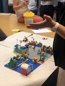A person gestures towards a colorful Lego model on a table. Several people are gathered around the table, one with arms folded and others sitting. The model includes miniature buildings, vehicles, and trees, resembling a small town or scene.