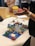 A person gestures towards a colorful Lego model on a table. Several people are gathered around the table, one with arms folded and others sitting. The model includes miniature buildings, vehicles, and trees, resembling a small town or scene.