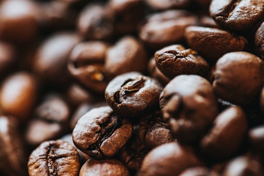 An up-close view of roasted coffee beans displaying their rich, dark brown texture and oily surface. The focus is on a cluster of beans with some slightly blurred in the background, emphasizing their natural, organic appearance.