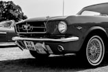 A classic car with a shiny exterior is prominently displayed. The vehicle features iconic detailing such as a front grille emblem and retro wheels. Nearby, another vintage car can be partially seen. The image is in black and white, highlighting the car's sleek lines and design.