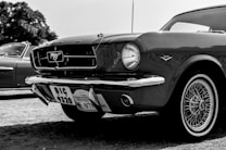 A classic car with a shiny exterior is prominently displayed. The vehicle features iconic detailing such as a front grille emblem and retro wheels. Nearby, another vintage car can be partially seen. The image is in black and white, highlighting the car's sleek lines and design.