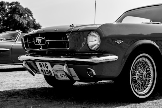 A classic car with a shiny exterior is prominently displayed. The vehicle features iconic detailing such as a front grille emblem and retro wheels. Nearby, another vintage car can be partially seen. The image is in black and white, highlighting the car's sleek lines and design.